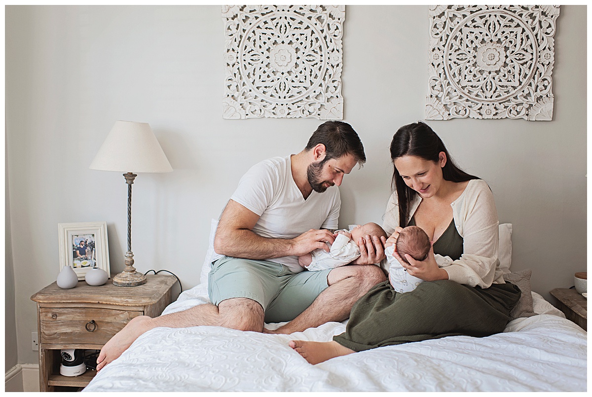 A mother and father sit together on a bed with their newborn baby twin boys
