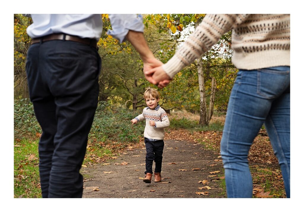 Boy running to his parents who hold hands in Richmond Park for an Autumn Family Photoshoot 