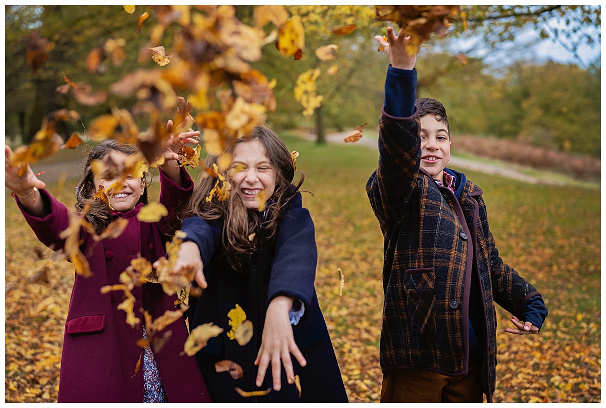 Three children throwing leaves at each other during an autumn family photo shoot in Richmond Park, south west London