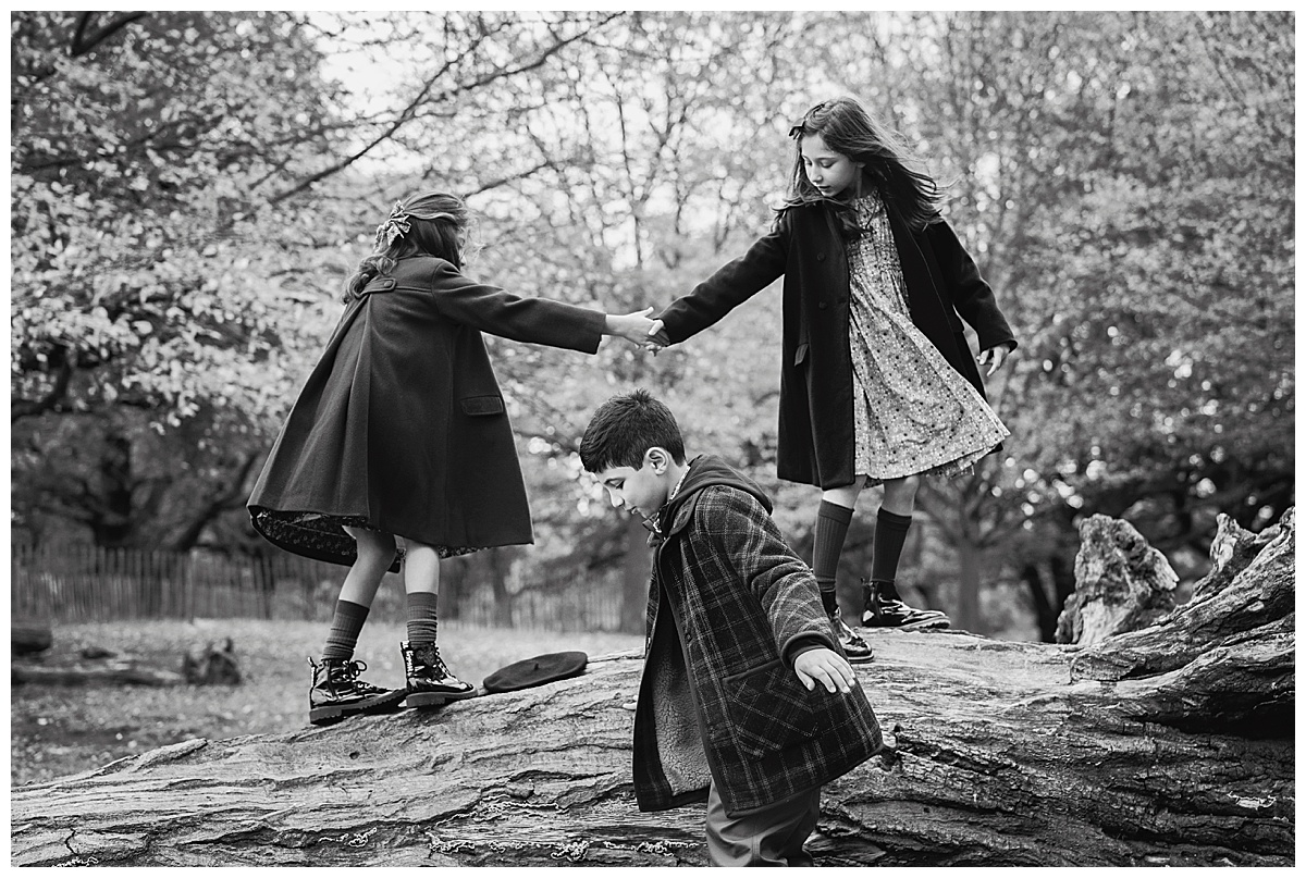 Black and white image of siblings climbing on logs in Richmond Park in Richmond upon Thames during a family photo shoot