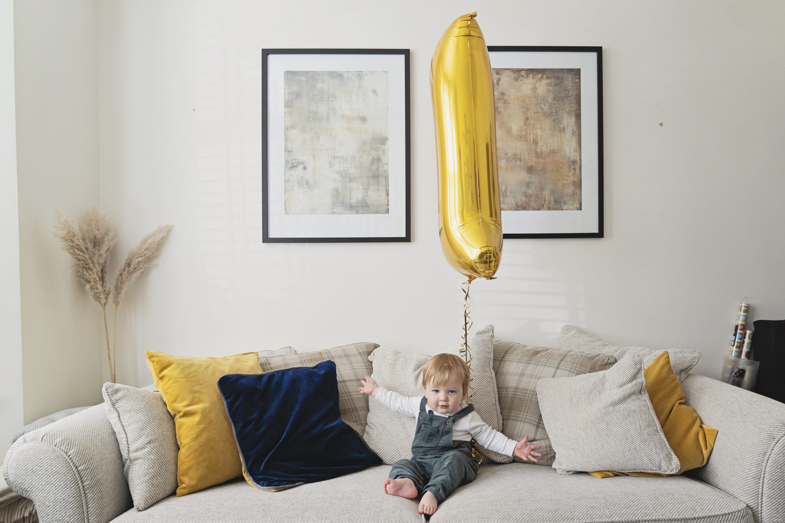 One year old baby boy sits on a sofa with a yellow number one helium balloon as part of a photo shoot to celebrate his first birthday.