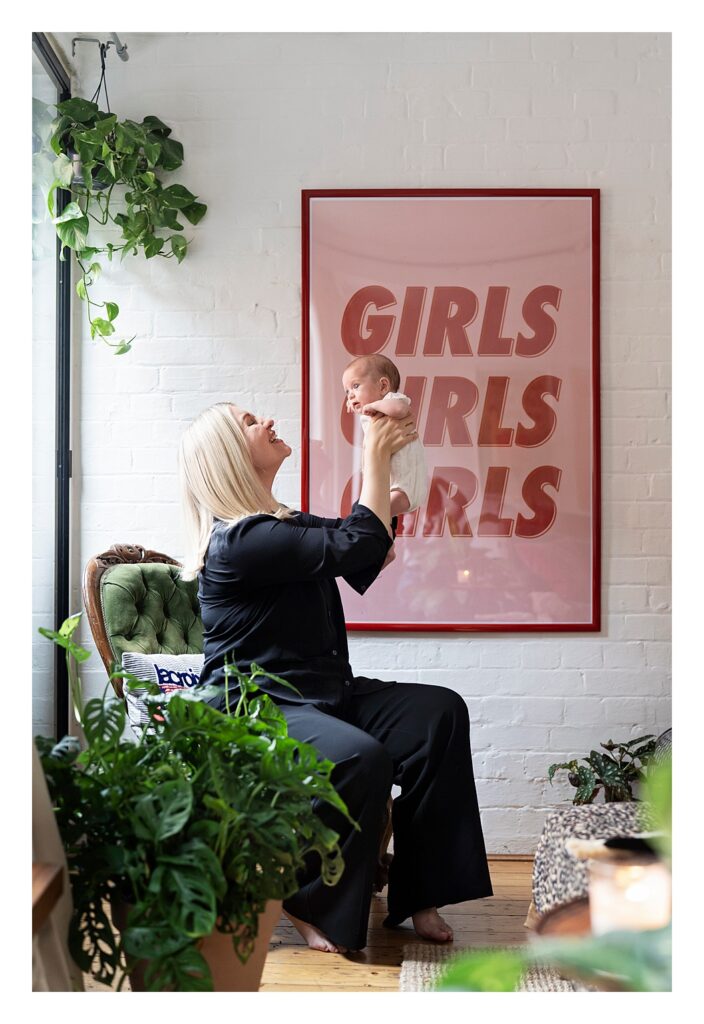 Mother holds her baby girl up against a framed print that reads "girls, girls, girls".