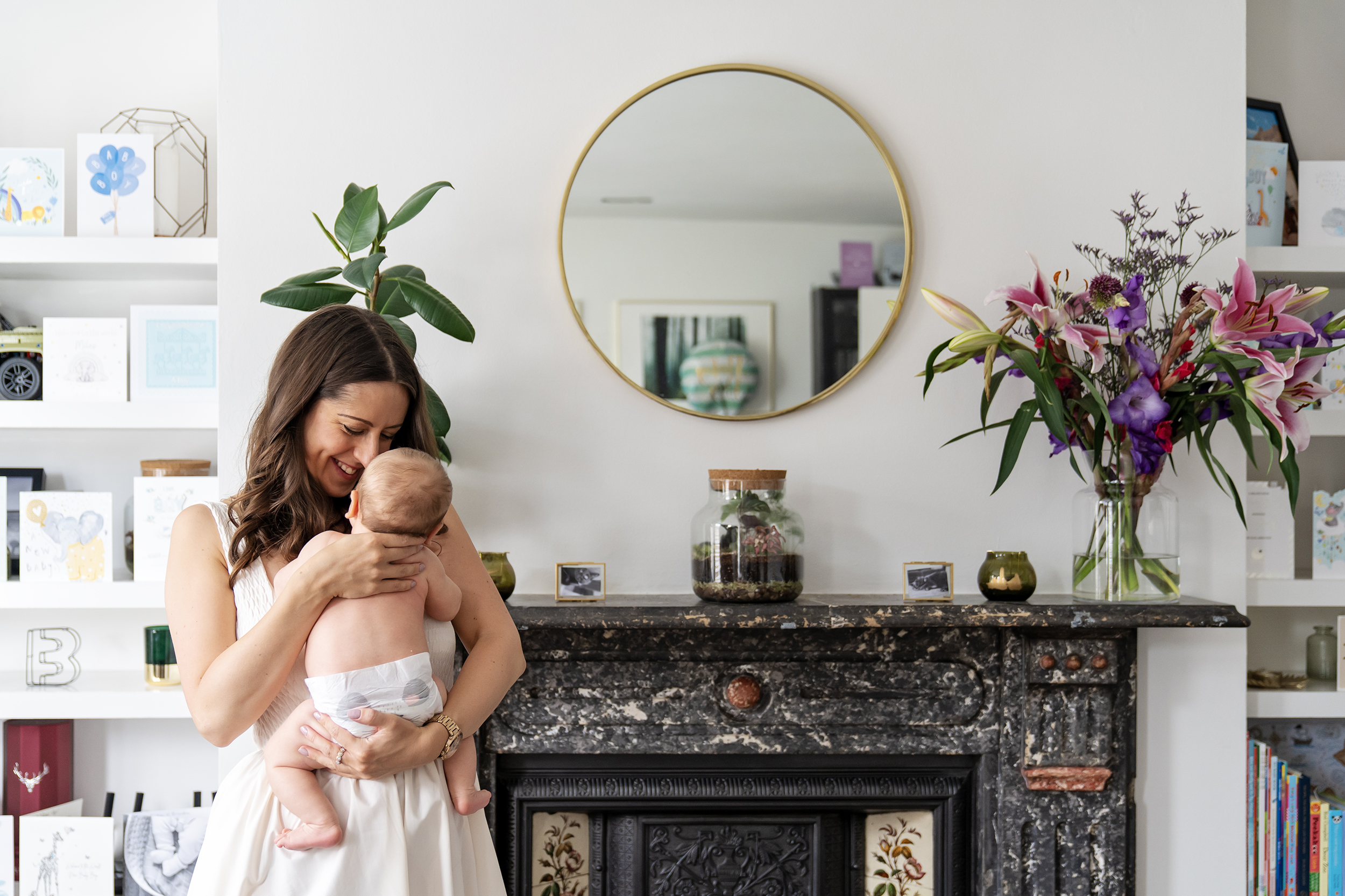 Mother smiles and cuddles are newborn baby bot close infant of her fireplace during an at home newborn photo shoot in London
