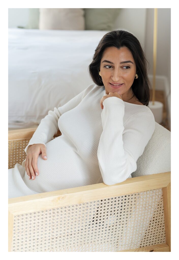 28-week pregnant woman sits in a rocking chair stroking her baby bump. She wears a cream dress for a maternity photo shoot at her home in London.