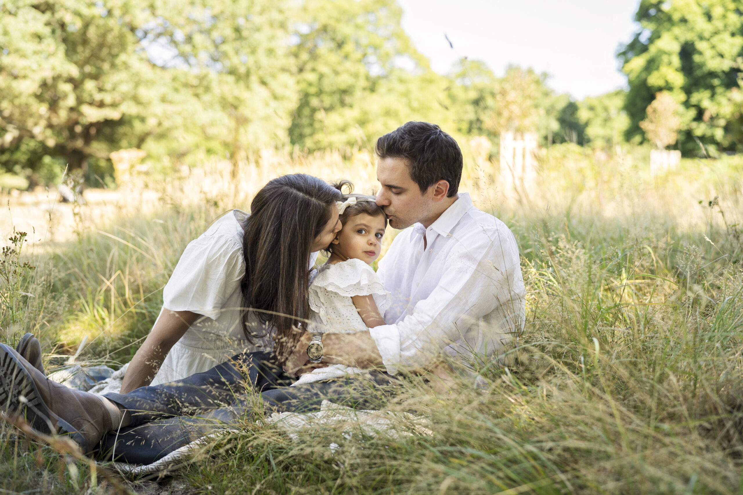 A mother and father kiss their little girl as they sit in the summer grasses at London's Richmond Park.