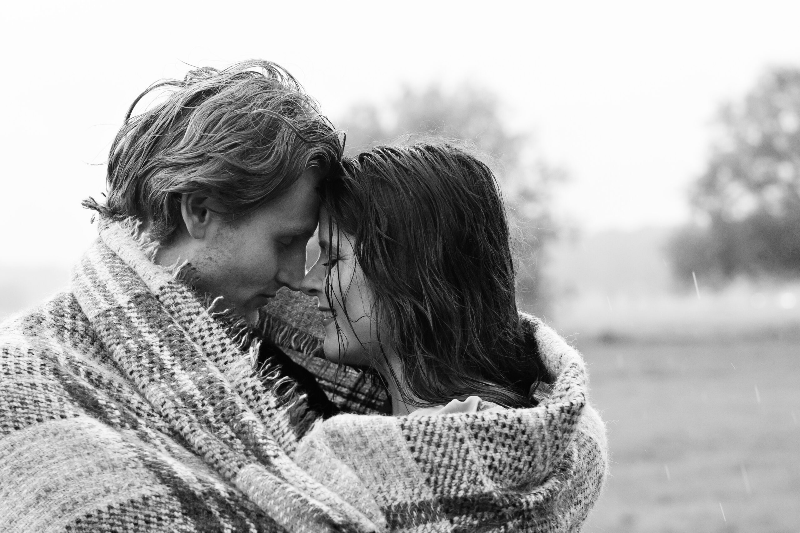 A couple stand eye to eye ,wrapped in a blanket during a rainstorm.